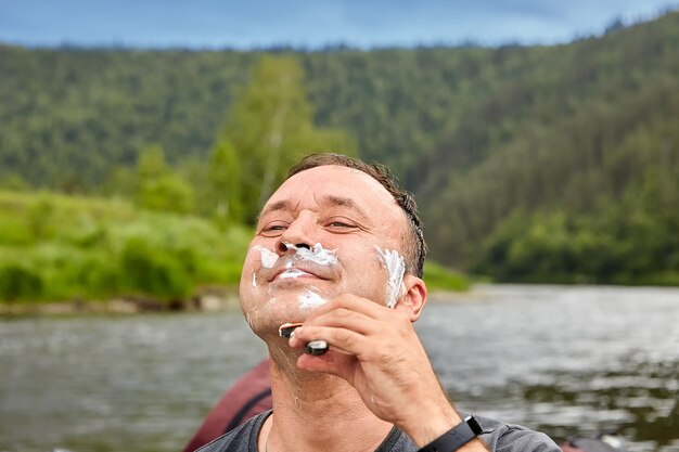 Homem sorridente faz a barba na natureza usando espuma de barbear e navalha.