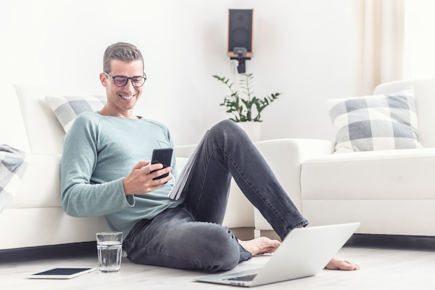 Homem sorridente mensagens de texto em seu telefone celular durante o horário de seu escritório em casa em casa, sentado no chão da sala de estar.