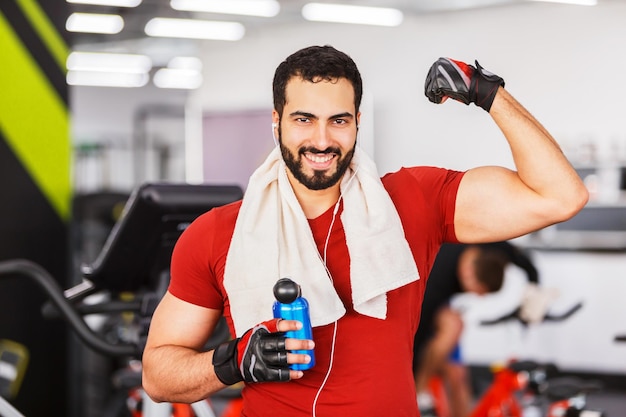 Homem sorridente musculoso barbudo usa camiseta vermelha mostra bíceps no descanso trabalhador do ginásio