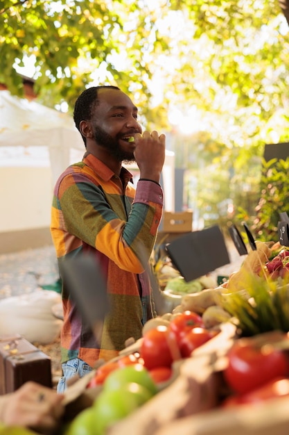 Homem sorridente provando maçã antes de comprar produtos biológicos, comprando produtos orgânicos naturais no mercado de agricultores. Cliente jovem desfrutando de comida com degustação de produtos locais frescos.