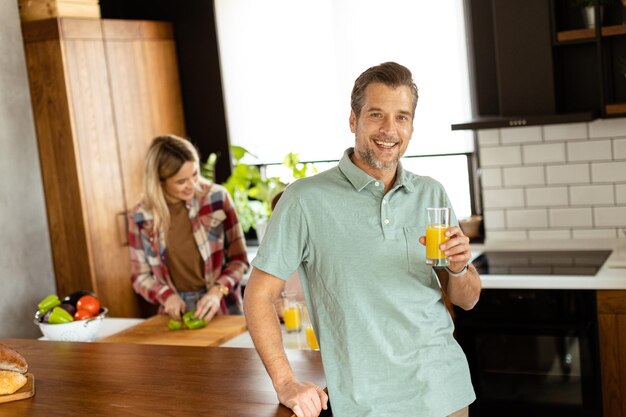 Homem sorridente segura um copo de suco de laranja enquanto duas mulheres cozinham em uma cozinha brilhante