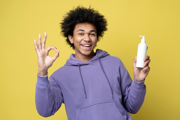 Homem sorridente segurando o frasco de xampu de cabelo, mostrando sinal ok isolado em fundo amarelo. Cuidado capilar