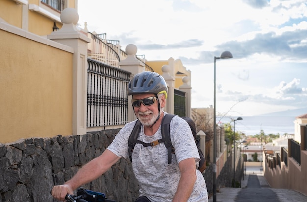 Homem sorridente sênior com capacete esporte e óculos de sol, andar de bicicleta com sua bicicleta elétrica em uma viela íngreme ao pôr do sol. Horizonte sobre a água em segundo plano