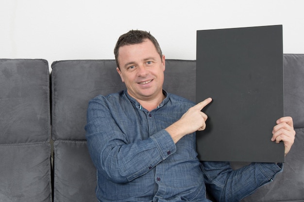 Homem sorridente sentado no sofá segurando uma placa preta em branco