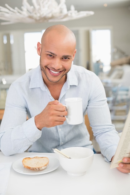 Foto homem sorridente tomando café enquanto lê o jornal em casa