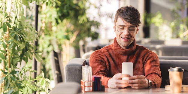 Homem sorridente usando smartphone perto de copo descartável no café