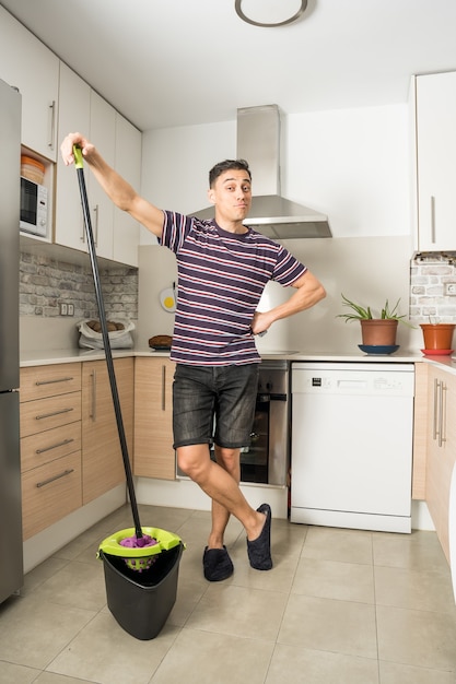 Homem sorridente, vestindo roupas casuais e chinelos, limpando o chão da cozinha. corpo todo.