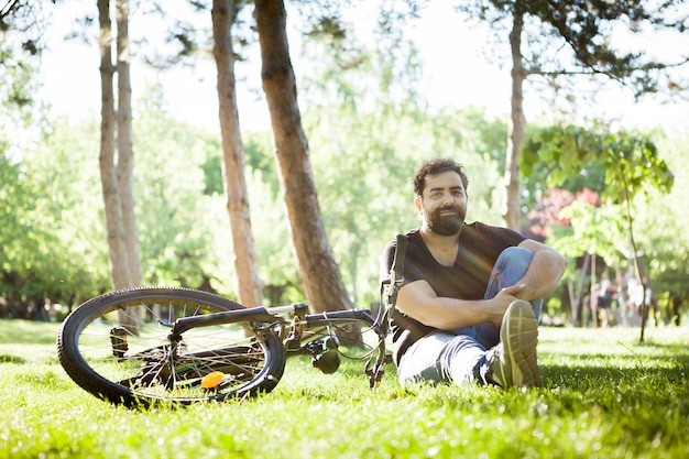 Homem sorrindo para a câmera sentado no chão na grama em um parque ao lado de sua bicicleta