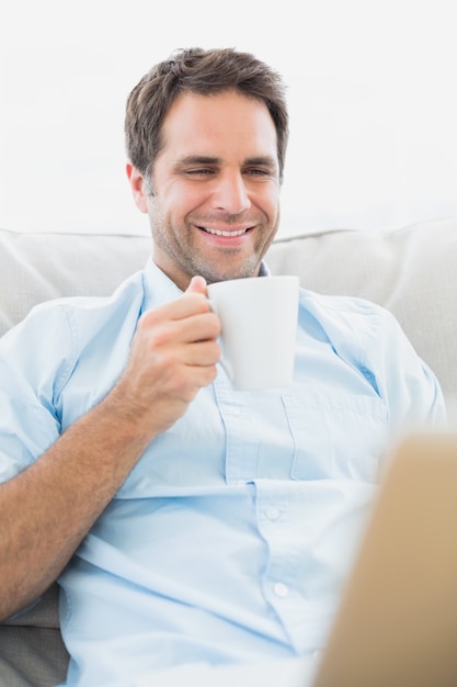 Homem sorrindo usando um laptop sentado no sofá com um café