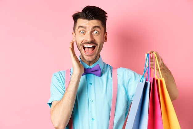 Homem surpreso e feliz gritando de alegria e segurando sacolas de compras, o comprador vendo descontos, em pé sobre rosa.