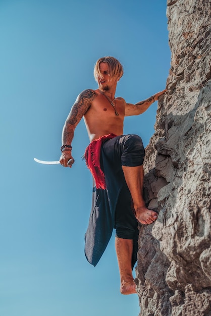 Homem tatuado maduro segurando a espada pronta para lutar e subir na rocha com fundo azul