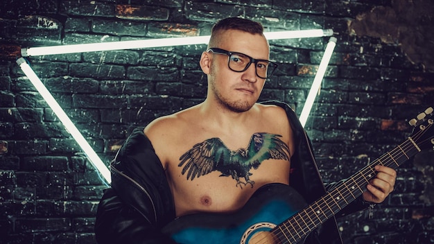 Homem tatuado tocando guitarra perto de parede iluminada Cara elegante com tatuagem de pássaro no peito olhando para longe e tocando guitarra em pé contra a parede de tijolos gasto com iluminação triângulo