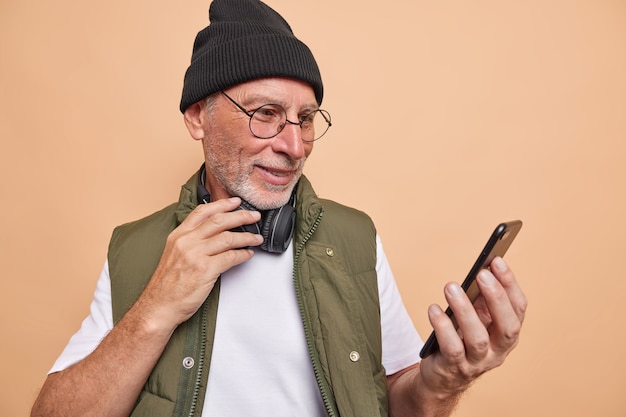 homem telefone usuário navega internet faz videochamada usa fones de ouvido estéreo usa boné camiseta e colete óculos transparentes poses em marrom