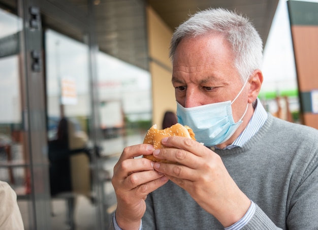 Homem tentando comer seu hambúrguer de fast food enquanto usava uma máscara