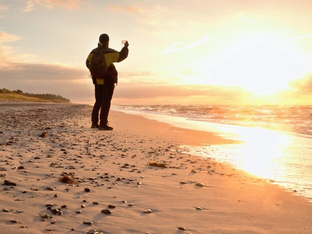 Foto homem tira foto de um caminhante marítimo à noite com mochila e telefone na mão ao longo da praia em estilo vintage