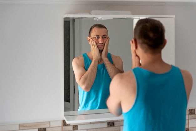 Homem tocando sua barba e rosto Homem sonolento no banheiro olha para si mesmo no espelho Rotina matinal