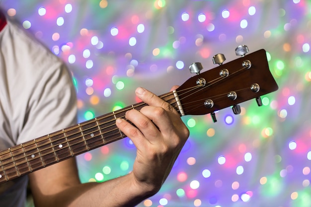 Homem tocando violão. Closeup dedos no pescoço da guitarra contra luzes desfocadas de bokeh de Natal no fundo.