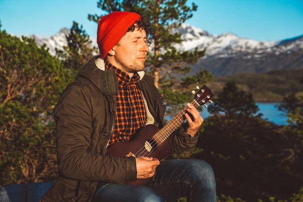 Homem tocando violão no fundo de montanhas, florestas e lagos, usa uma camisa