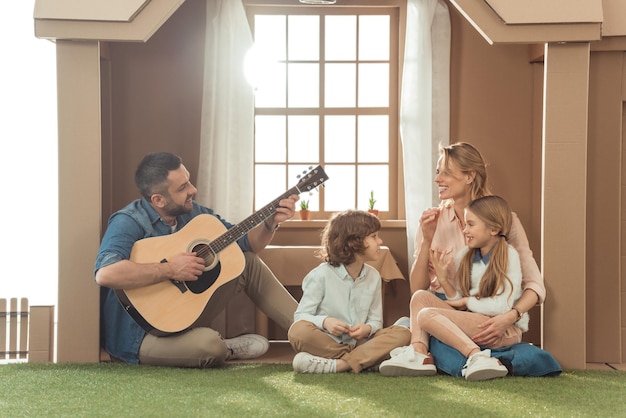 homem tocando violão para sua família na nova casa de papelão