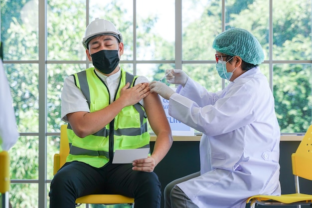 Homem trabalhador sentado para tomar a vacina covid, com a enfermeira injetando vacina para obter imunidade para proteger o vírus. Vacinação para Trabalhadores Essenciais em Clínica de Saúde em Fábrica Industrial.