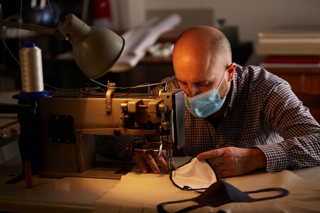 Homem trabalhando com máquina de costura, fazendo máscara facial caseira para prevenir e impedir a propagação do vírus corona