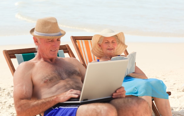 Homem trabalhando em seu laptop enquanto sua esposa lê na praia