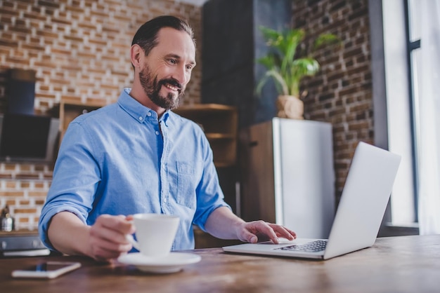 Homem trabalhando no laptop e tomando café na cozinha pela manhã