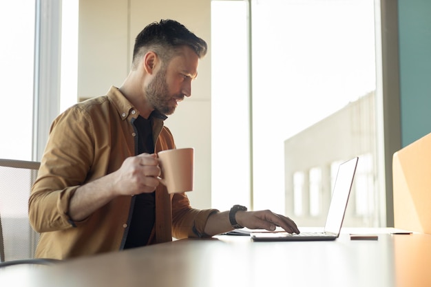 Homem trabalhando no laptop tomando café na vista lateral do escritório