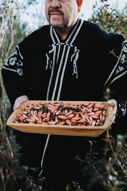 Homem tradicional usando um poncho de lã, segurando uma calha de madeira rústica com pinhões de araucária chilena