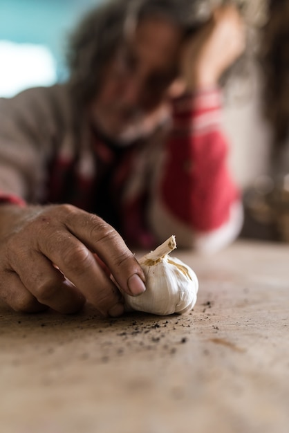 Homem triste olhando para o bulbo de alho, segurando-o com as mãos sujas enquanto se senta em uma mesa de madeira rústica.