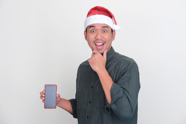 Homem usando chapéu de natal mostrando expressão espantada enquanto segura a tela do telefone em branco