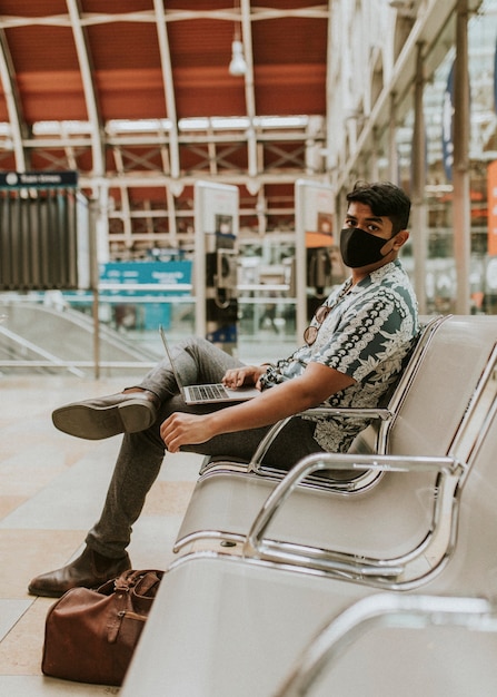 Homem usando máscara usando um laptop em uma estação de trem no novo normal