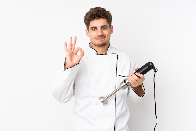 Homem usando o liquidificador isolado na parede branca, mostrando um sinal de ok com os dedos