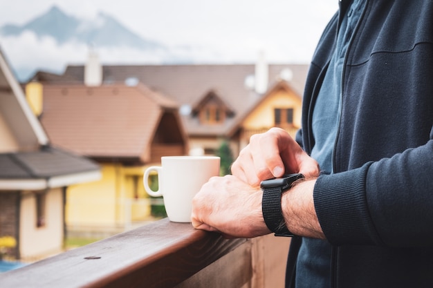 Homem usando seu relógio inteligente na varanda e tomando café com uma vista incrível das montanhas