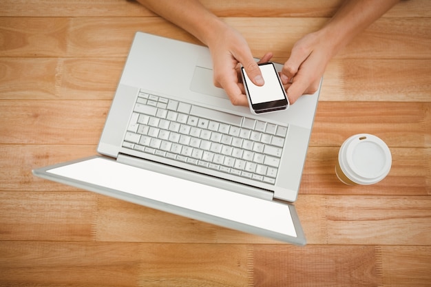 Homem usando smartphone enquanto laptop e copo descartable na mesa