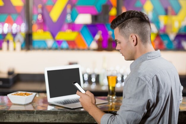 Homem usando telefone celular com o laptop na mesa no balcão de bar