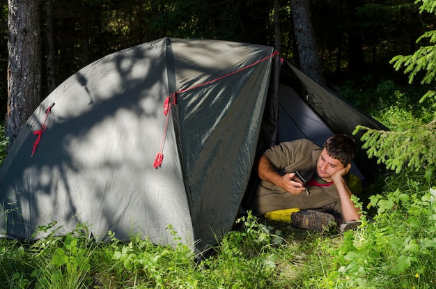 Foto homem usando telefone celular enquanto está deitado na tenda na floresta
