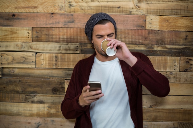 Homem usando telefone celular enquanto toma um café contra a parede de madeira