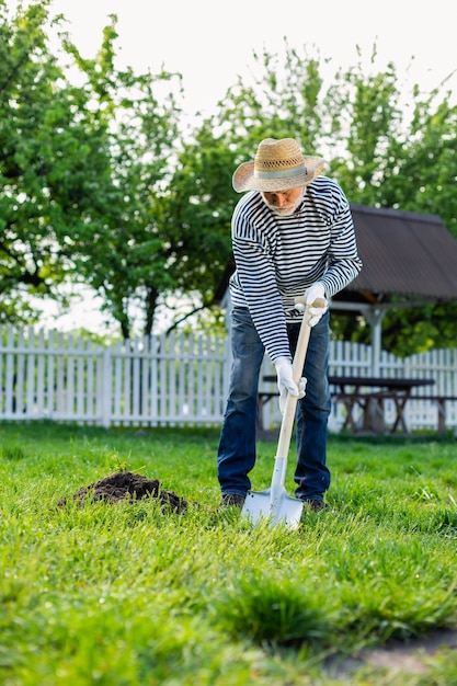 Homem usando uma pá. Homem com chapéu de palha usando luvas brancas enquanto usa uma pá no jardim perto de sua casa de verão