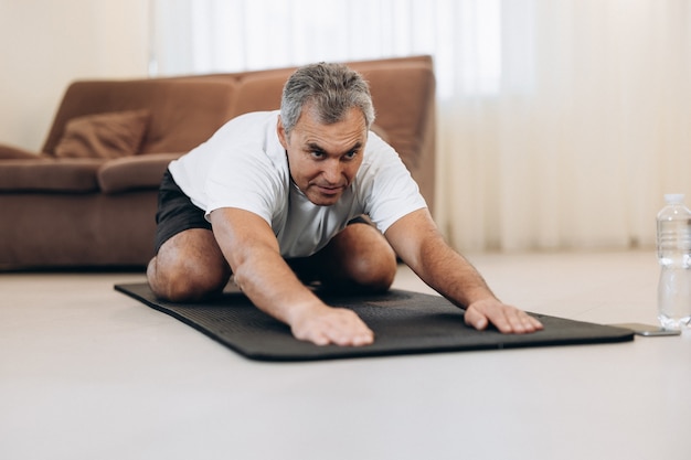 Homem velho em roupas esportivas deitado em colchonetes fazendo asana infantil, exercícios para acalmar o corpo e a mente, esticando as mãos para a frente