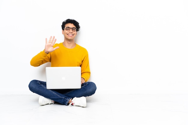 Homem venezuelano sentado no chão com laptop contando cinco com os dedos