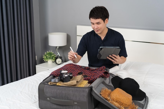 Homem verifica uma lista de coisas com um tablet preparar e embalar roupas na mala em uma cama em casa planejando férias de viagem