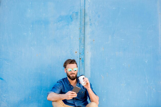 Homem vestido casual com camisa azul sentado com telefone e café para ir no fundo da parede azul. Plano amplo com espaço de cópia