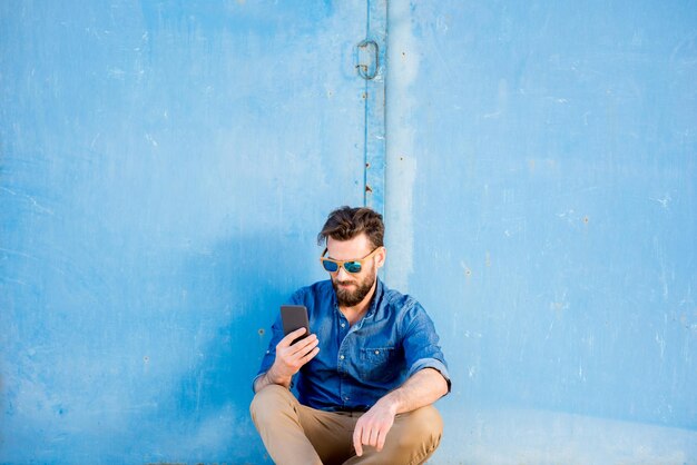 Homem vestido casual com camisa azul sentado com telefone no fundo da parede azul. Plano amplo com espaço de cópia