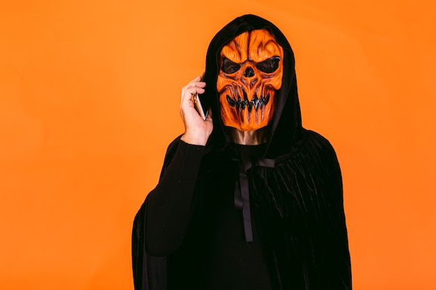 Homem vestido com máscara de abóbora de látex e capa de veludo fala em seu telefone celular. Halloween e dias do conceito morto.