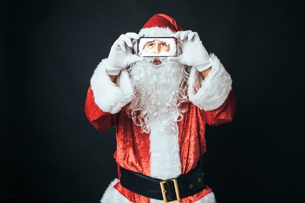 Homem vestido de Papai Noel com um celular nas mãos, tirando uma selfie, sobre fundo preto. Conceito de Natal, Papai Noel, presentes, celebração.