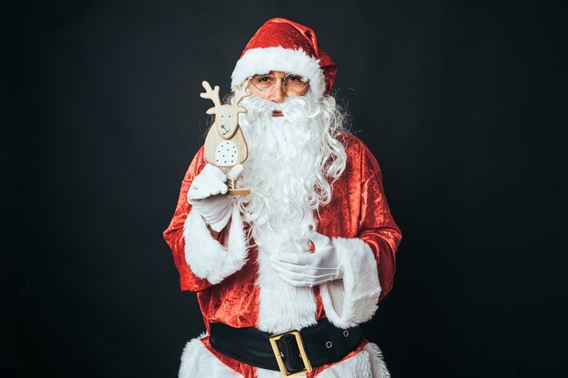 Homem vestido de Papai Noel segurando a figura de uma rena de madeira, sobre fundo preto. Conceito de Natal, Papai Noel, presentes, celebração.