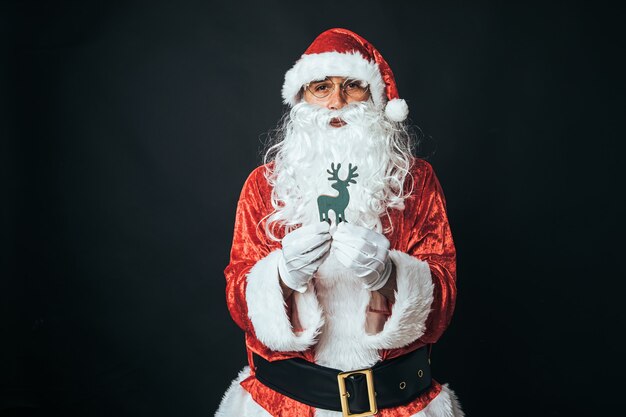 Homem vestido de papai noel segurando uma figura de madeira de uma rena, sobre fundo preto. conceito de natal, papai noel, presentes, celebração.