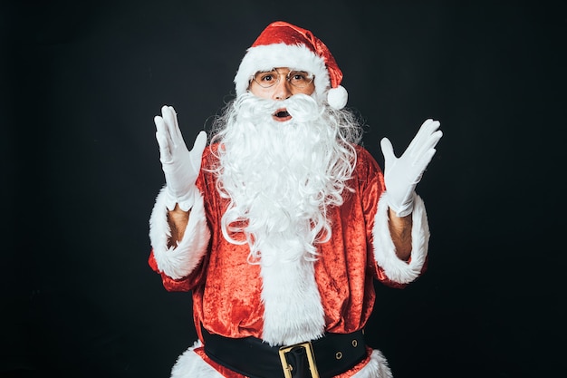 Homem vestido de Papai Noel surpreso, com as mãos levantadas, sobre fundo preto. Conceito de Natal, Papai Noel, presentes, celebração.