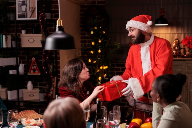 Homem vestindo fantasia de papai noel dando presente de natal para mulher, esposa feliz recebendo presente do marido. Celebração da noite de Natal com grande família na mesa de jantar festiva, saudação de férias de inverno